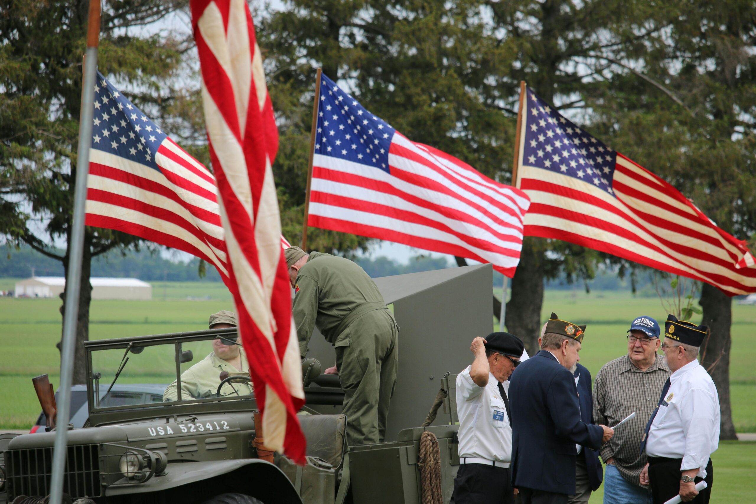 Group of veterans and soldiers by military vehicle with American flags in the background, symbolizing patriotism.